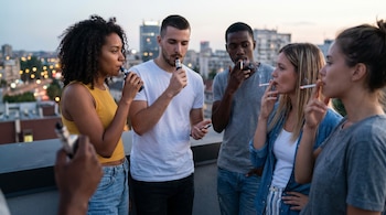 Un grupo de jóvenes adultos en una azotea urbana al atardecer; algunos fuman cigarrillos mientras otros usan vapeadores con la ciudad de fondo.