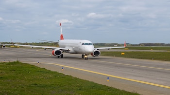 Un avión comercial blanco con detalles rojos en la cola y motores avanza sobre una pista de rodaje de asfalto en un día nublado
