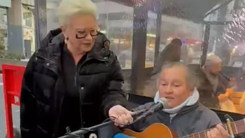 Uma mulher loira platinada e óculos (Carmen Barbieri) sorri e segura um microfone para um guitarrista de rua.