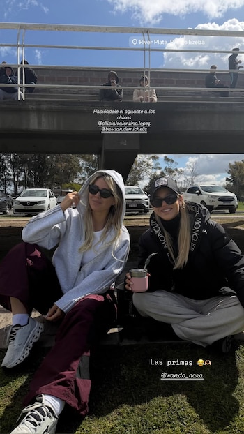 Wanda Nara y Evangelina Anderson sentadas sobre el césped con gafas de sol frente a unas tribunas y varios vehículos estacionados