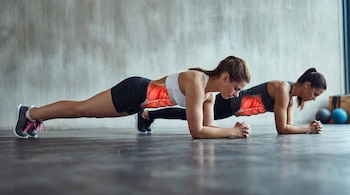Dos mujeres jóvenes, vestidas con ropa deportiva, en posición de plancha sobre el suelo de un gimnasio; sus músculos abdominales están resaltados en rojo.