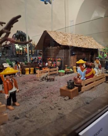 Diorama de PLAYMOBIL con figuras humanas, cerdos, gallina, palomas, una cabaña de madera con techo de paja, fardos de heno y cestas de vegetales