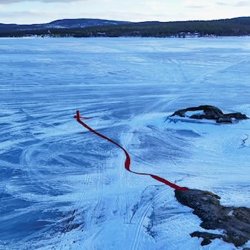 Vista aérea de una gran superficie de Sea-M congelado cubierta de hielo y nieve, con un lazo rojo brillante que se extiende desde una roca oscura hacia un punto en forma de cruz en el hielo. Se aprecian montañas cubiertas de árboles y un cielo nublado al fondo