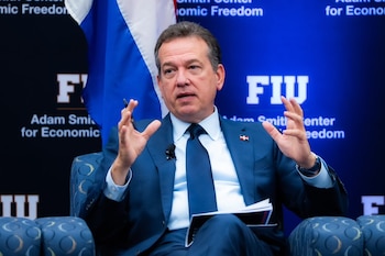 Hombre en traje y corbata, sentado, gesticula con ambas manos. Lleva pin de bandera dominicana y micrófono. Fondo azul con texto FIU y Adam Smith Center