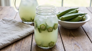 Jarra de vidrio y vaso con agua de okra, hielo y rodajas de okra; un recipiente blanco con vainas de okra enteras, mantel y mesa de madera.