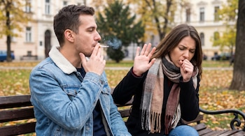 Un hombre fuma un cigarrillo en un banco de madera. A su derecha, una mujer tose y aleja el humo con su mano, sentados en un parque con hojas caídas.