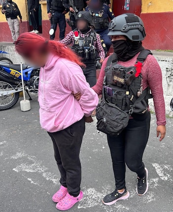 Vista de una mujer de cabello rojo y sudadera rosa siendo escoltada por una oficial de policía con casco y chaleco negro que dice "EXPLOSIVOS" en la calle