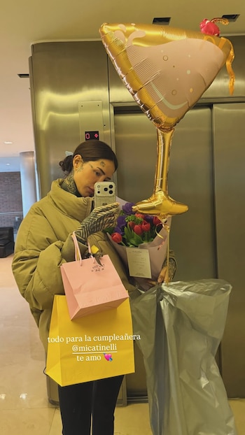 Cande Tinelli se toma una foto sosteniendo un ramo de flores, bolsas de compras y un globo dorado con forma de copa frente a un ascensor