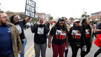Grupo de personas protestando en una calle. Algunos gritan. Se ve un cartel 'BLACK VOTERS MATTER' y camisetas con 'I'M BLACK & I WANT $15'