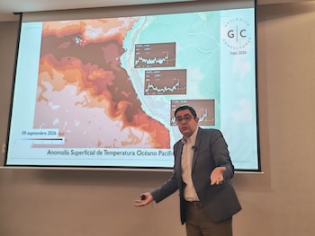 Un hombre habla con las manos abiertas frente a una pantalla proyectada que muestra un mapa térmico del océano Pacífico y tres gráficos de líneas