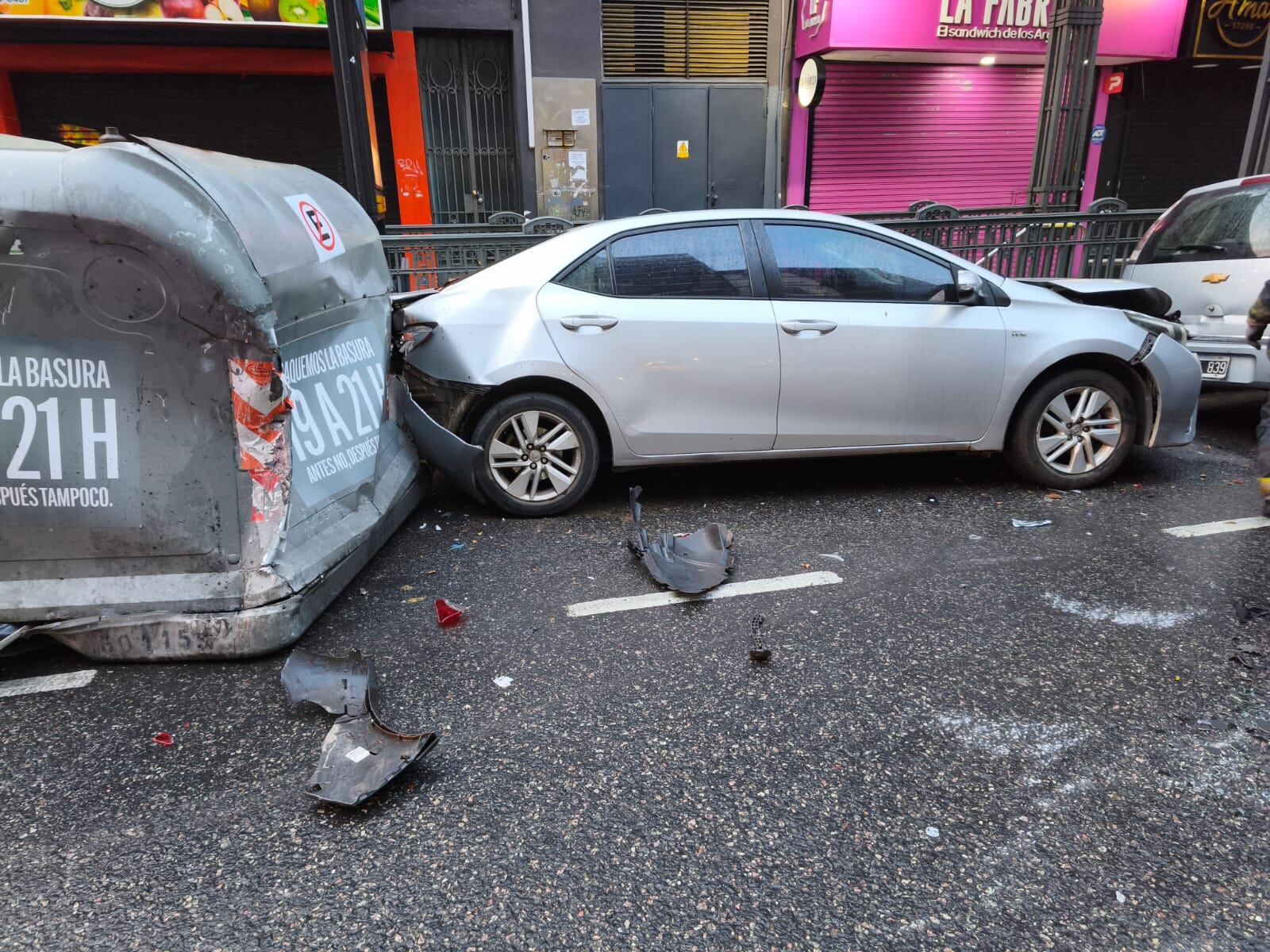 Un Toyota sedan plateado presenta daños en la carrocería tras impactar contra un contenedor de residuos en una calle urbana, con fragmentos del vehículo esparcidos en el asfalto.