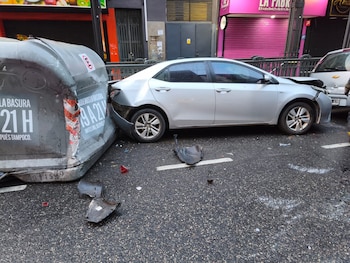 Toyota sedan plateado con daños en frontal y parte trasera, impactado contra contenedor de residuos gris. Fragmentos de coche en el asfalto mojado. Edificios urbanos