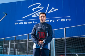 Hombre con chaqueta azul, sostiene raqueta de pádel negra y pelota de tenis amarilla. Mira a la derecha. Detrás, edificio grande y elegante bajo cielo azul