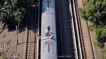 Vista aérea de un tren de pasajeros en movimiento con cuatro personas sobre su techo, circulando entre vías y zonas de árboles verdes.
