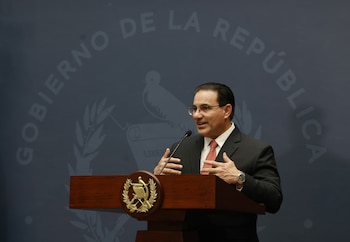 Hombre de traje y gafas pronuncia un discurso detrás de un podio de madera con un emblema dorado. El fondo es azul oscuro con texto "Gobierno de la República"