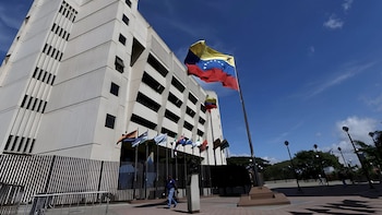 Una bandera de Venezuela ondea en un mástil frente a un edificio blanco de varios pisos con otras banderas pequeñas a la entrada