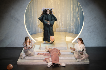 Escena teatral con cinco mujeres: una de pie en abrigo negro, una tumbada, dos sentadas. Fondo gris con un gran círculo luminoso y cortinas translúcidas. Dos globos en el suelo