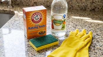 Un envase de bicarbonato de sodio y una botella de vinagre blanco, junto a una esponja y guantes amarillos sobre una encimera de granito.