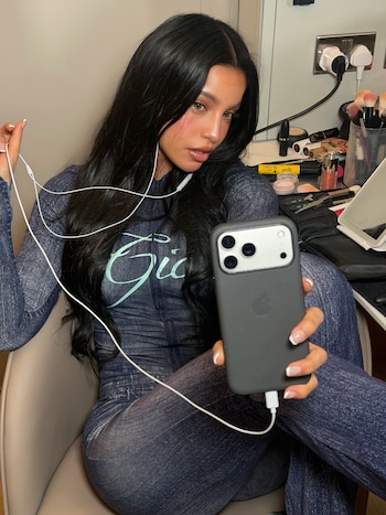 Una mujer de largo cabello oscuro sostiene un teléfono móvil y unos auriculares mientras posa frente a una mesa con artículos de maquillaje