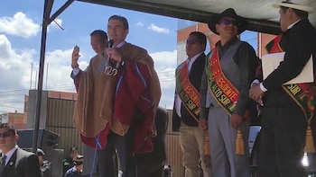Un hombre en poncho marrón y camisa clara habla en un micrófono, con su mano derecha levantada, flanqueado por otros hombres, algunos con bandas presidenciales