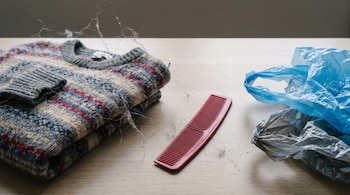 Un suéter de lana doblado con rayas grises, rojas y azules, un peine de plástico rojo, una bolsa plástica azul y otra gris, con pelusas y fibras.