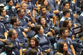 Grupo de pessoas sentadas em uniformes azuis escuros e azuis claros com insígnias. Um homem está segurando um telefone e o outro está segurando óculos. Há sinais indicando