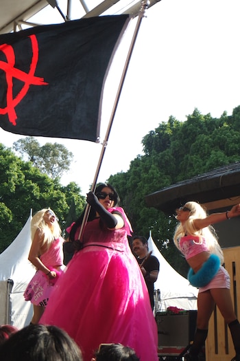 Una persona con vestido rosa sostiene una bandera negra con un símbolo anarquista junto a dos bailarinas con pelucas rubias en un escenario