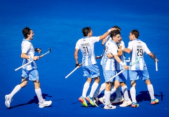 Jugadores de hockey con uniforme celeste y blanco se abrazan sosteniendo sus palos sobre una cancha de color azul