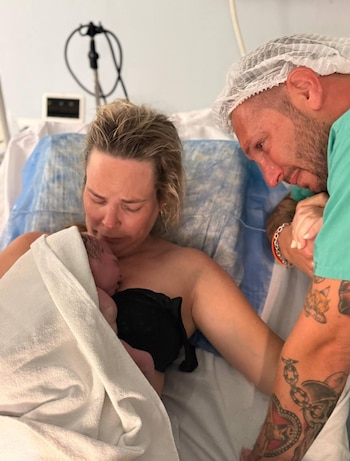 Mujer con el torso desnudo sostiene un recién nacido envuelto en toalla blanca. Un hombre con gorro y uniforme médico mira emocionado en sala de hospital