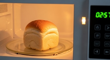 Un pan cuadrado de color dorado y blanco se encuentra sobre un plato de cristal dentro de un microondas encendido, con la pantalla digital marcando el tiempo.