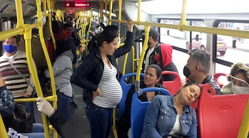 Una mujer embarazada se sostiene de un tubo en un bus TransMilenio lleno, con otras dos mujeres sentadas en sillas azules frente a ella; una duerme y la otra la mira.