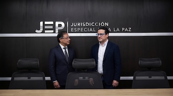 Dos hombres de traje de pie frente a una pared negra con el logo y el nombre de la Jurisdicción Especial para la Paz (JEP). Una mesa y sillas.