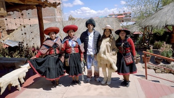 Cinco personas posan de pie al aire libre. Tres mujeres visten ropa tradicional andina con sombreros de ala ancha. Un hombre y una mujer joven visten ropa moderna