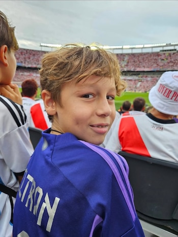 Primer plano de un niño sonriendo con pelo rubio y aparatos, vistiendo camiseta azul de fútbol con "BELTRON" en la espalda, sentado en un estadio con público