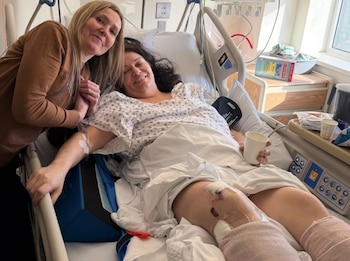 Una mujer en una cama de hospital con bata y vendajes en las rodillas sonríe, mientras otra mujer rubia la mira y le toma la mano