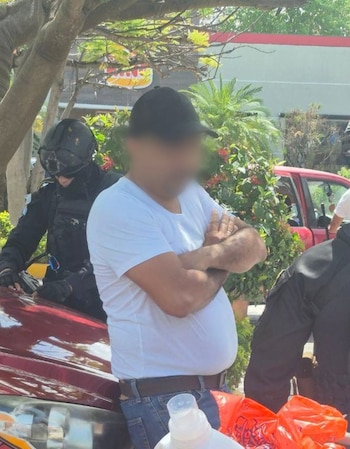 Un hombre con camiseta blanca y rostro difuminado permanece junto a agentes con equipo táctico y un vehículo de color rojo