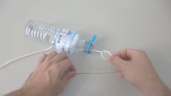 Dos manos sostienen una cuerda blanca con un nudo que atraviesa una botella de plástico transparente sobre una superficie clara