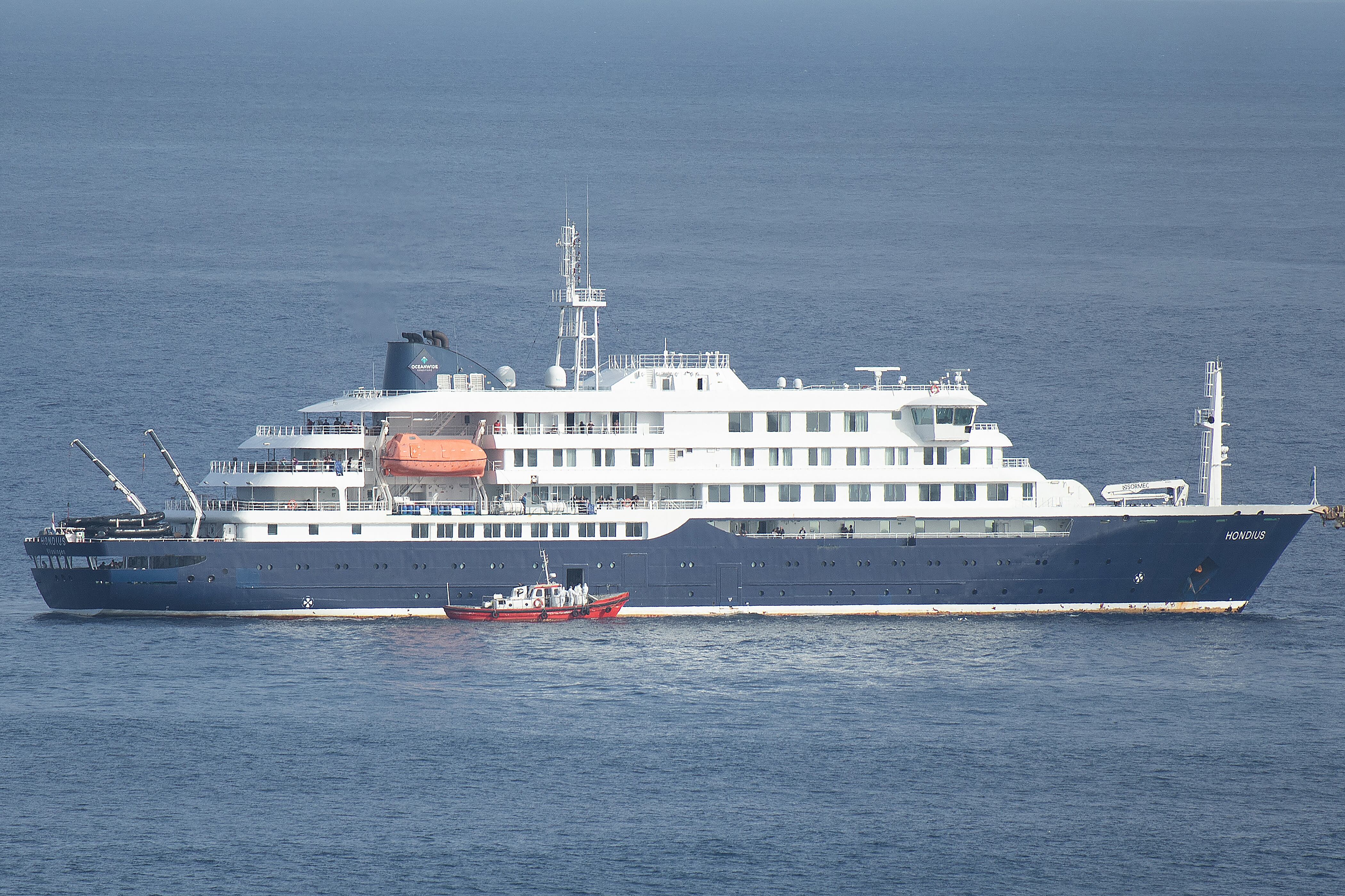 Una lancha ambulancia con tripulantes vestidos con trajes de protección se aproxima a la puerta de práctico en el costado de estribor del crucero MV Hondius, mientras permanece detenido frente al puerto de Praia, la capital de Cabo Verde, el 5 de mayo de 2026, mientras los pasajeros observan desde los puentes. (AFP)
