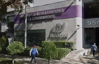 Fachada gris de las oficinas de Prodecon con un logo plateado y un letrero morado. Vegetación frondosa en primer plano y dos personas caminando cerca