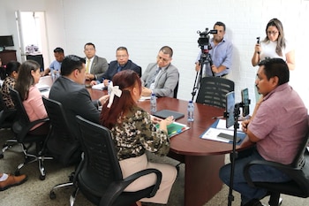 Grupo de personas en una sala de reuniones con paredes blancas, sentadas alrededor de una mesa de madera. Se observa la bandera de Guatemala y una pantalla