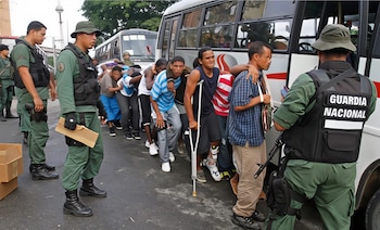 Hombres con uniforme verde militar y chaleco negro con la inscripción 'GUARDIA NACIONAL'. Fila de hombres civiles abordando un autobús, uno usa una muleta. Otros autobuses
