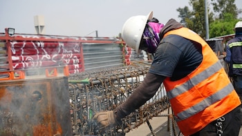 Un obrero con casco blanco y chaleco reflectante anaranjado trabaja sobre una estructura de acero en una obra
