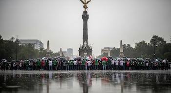 Multitud con paraguas y camisetas verdes, blancas, rojas se reúne frente al Ángel de la Independencia bajo lluvia intensa; reflejos en el suelo mojado.