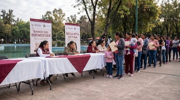 Madres hacen fila para recibir tarjetas de programas sociales en un parque, con mesas, banners oficiales y una niña junto a su madre.