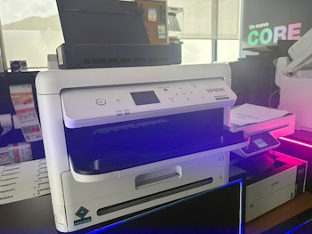 Impresora multifunción Epson blanca y negra. Unidad superior de escáner. Papeles en bandeja. Pantalla y botones de control. Iluminación led azul y magenta