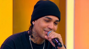 Primer plano de Youna con un gorro negro, pendiente, piercing nasal y tatuajes en el cuello, sosteniendo un micrófono frente a un fondo degradado naranja y amarillo.