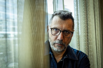 Retrato de hombre con gafas, barba canosa y chaqueta vaquera, mirando a cámara, con cortinas translúcidas a ambos lados y un fondo borroso