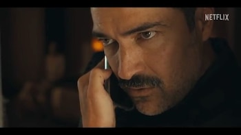 Primer plano de un hombre con bigote hablando por un teléfono celular. El logo de Netflix se ve en la esquina superior derecha