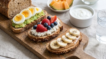 Rebanadas de pan integral con aguacate y huevo, requesón y frutos rojos, crema de cacahuete y plátano sobre una tabla de madera, junto a yogur, melón y agua.