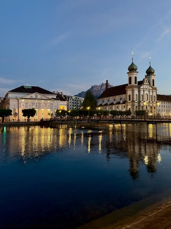 Vista nocturna de un río oscuro reflejando luces de edificios iluminados en sus orillas, incluyendo una iglesia con dos cúpulas verdes, y montañas claras al fondo